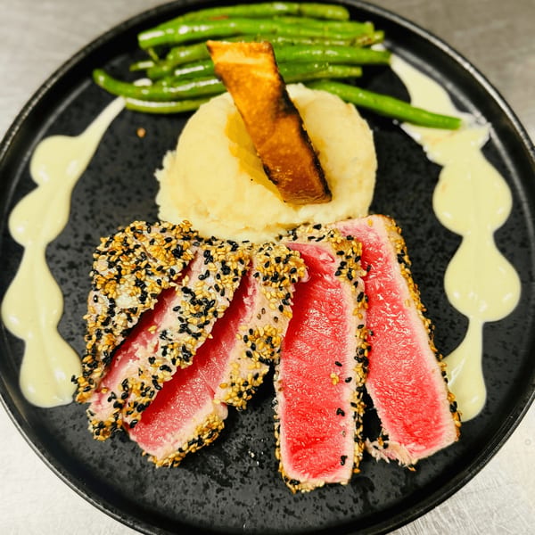 Seared tuna with sesame crust, mashed potatoes, green beans, and a creamy sauce on a black plate.