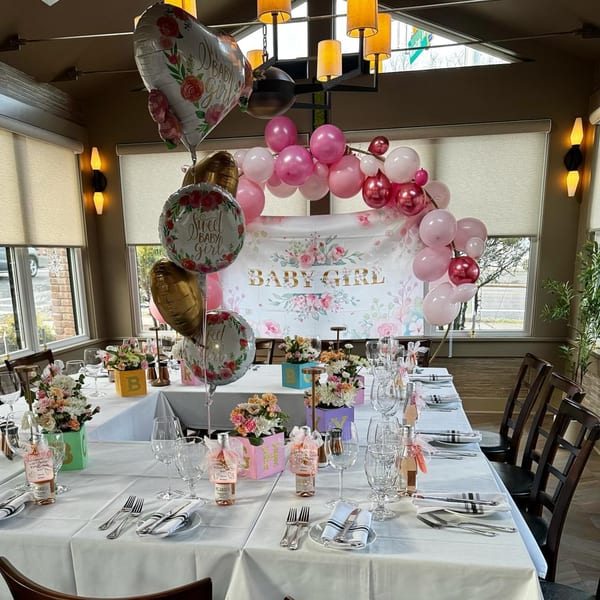 A restaurant dining room set up for a "Baby Girl" celebration with pink balloons and floral decorations.